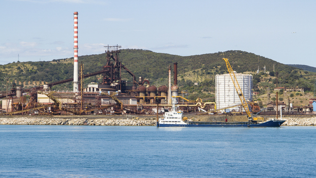 View of the industrial complex with its towering chimney and cranes contrasts with the calm blue sea and distant green hills Piombino Tuscany Italy. View of the industrial complex with its towering chimney and cranes contrasts with the calm blue sea and distant green hills Piombino Tuscany Italy.