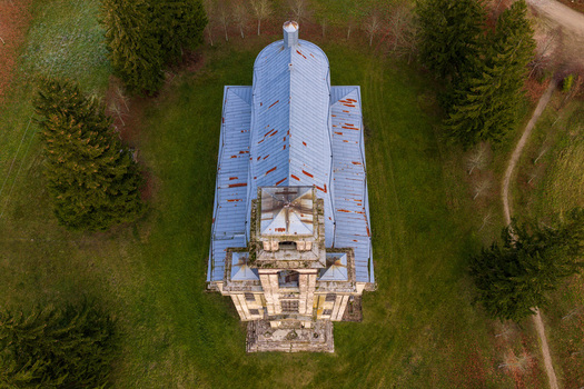 Aerial view of the historic Palendriai church with its weathered metal roof and stone tower surrounded by tall evergreen trees and green fields in Palendriai Siauliu apskritis Lithuania.