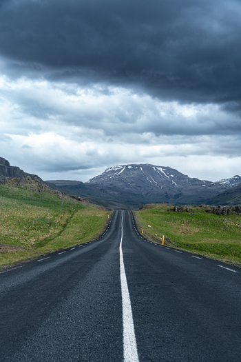 View of a long asphalt road leading towards the snow capped mountains of Hvalfjordur under a dramatic cloudy sky in Iceland. View of a long asphalt road leading towards the snow capped mountains of Hvalfjordur under a dramatic cloudy sky in Iceland.