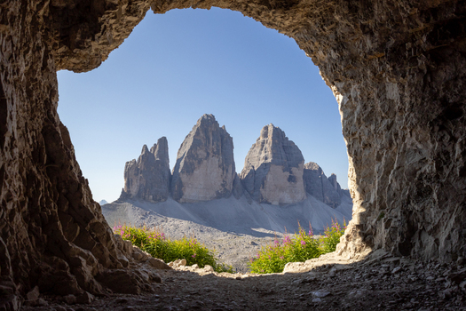 View of Tre Cime di Lavaredo framed by a natural rock cave opening with rugged stone textures vibrant wildflowers and clear blue sky in Auronzo di Cadore Veneto Italy.