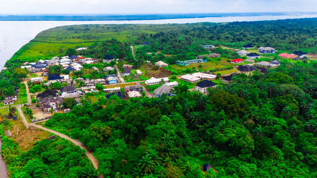 Aerial view of a settlement nestled within dense tropical vegetation near a wide river featuring houses with varied roof colors and winding paths in Minima Rivers Nigeria.