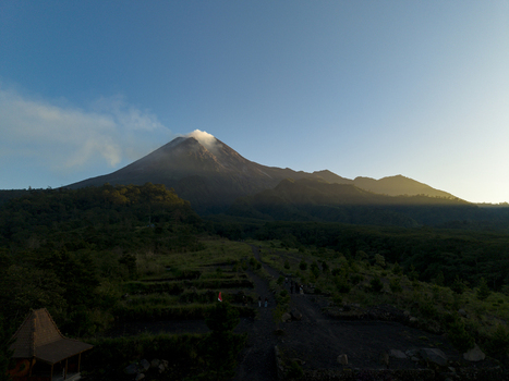 Aerial view of Bunker Kaliadem Merapi and the smoking peak of Mount Merapi volcano surrounded by lush green forests and a winding path Kecamatan Pakem Daerah Istimewa Yogya Indonesia. Aerial view of Bunker Kaliadem Merapi and the smoking peak of Mount Merapi volcano surrounded by lush green forests and a winding path Kecamatan Pakem Daerah Istimewa Yogya Indonesia.