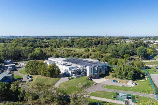 Aerial view of a modern data center facility featuring solar panels on the roof and external cooling units surrounded by lush green trees and open fields in the United Kingdom.