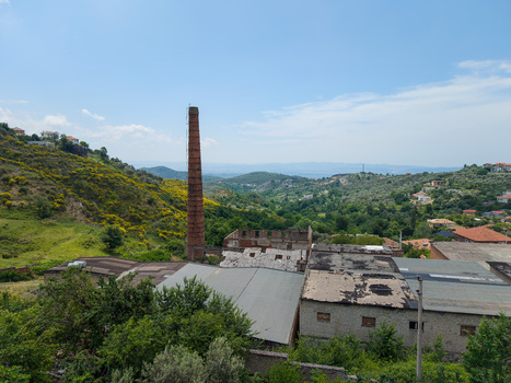 Aerial view of an abandoned industrial site with a tall brick chimney and weathered rooftops among green hills and yellow wildflowers in Kruje Durres County Albania.
