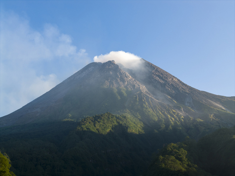 Aerial view of Mount Merapi volcano with smoke rising from its crater and lush green forests on the slopes under a clear blue sky Kecamatan Pakem Daerah Istimewa Yogya Indonesia. Aerial view of Mount Merapi volcano with smoke rising from its crater and lush green forests on the slopes under a clear blue sky Kecamatan Pakem Daerah Istimewa Yogya Indonesia.