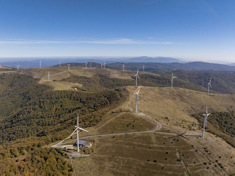 Aerial view of a wind farm with multiple white wind turbines situated on rolling hills covered with autumn forests under a clear blue sky in Bulgaria. Aerial view of a wind farm with multiple white wind turbines situated on rolling hills covered with autumn forests under a clear blue sky in Bulgaria.
