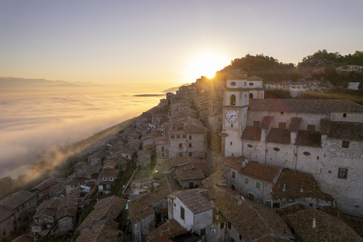 Aerial view of the historic town of Artena perched on a hillside above a sea of clouds at sunrise featuring stone rooftops and a clock tower in Artena Lazio Italy.