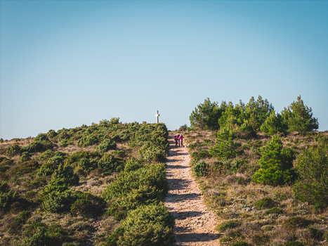 Aerial view of a winding path leading up a grassy hill towards a white cross framed by lush greenery under a clear sky Istria County Croatia.
