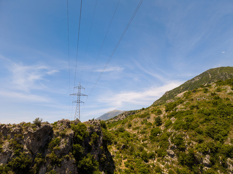 Aerial view of a high voltage power line tower standing on a rugged mountain ridge with rocky cliffs and green vegetation under a clear blue sky in Dhermi Vlore County Albania.