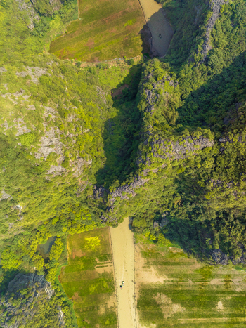 Aerial view of a winding river flowing through dramatic limestone karst mountains covered in lush green vegetation with small agricultural fields and a boat on the water in Vietnam. Aerial view of a winding river flowing through dramatic limestone karst mountains covered in lush green vegetation with small agricultural fields and a boat on the water in Vietnam.