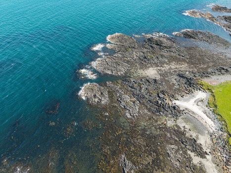 Aerial view of a rocky coastline with dark jagged stones meeting the deep blue sea and a patch of green grass in Donaghadee Northern Ireland United Kingdom. Aerial view of a rocky coastline with dark jagged stones meeting the deep blue sea and a patch of green grass in Donaghadee Northern Ireland United Kingdom.