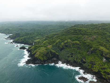 Aerial view of Pengilon Hill where turquoise waters meet the rugged coastline fringed by verdant forests under a soft overcast sky Gunungkidul Regency Indonesia. Aerial view of Pengilon Hill where turquoise waters meet the rugged coastline fringed by verdant forests under a soft overcast sky Gunungkidul Regency Indonesia.