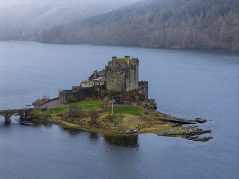 Aerial view of Eilean Donan Castle a historic stone fortress on a small island with a bridge surrounded by calm water and misty forested hills in Dornie Scotland United Kingdom.