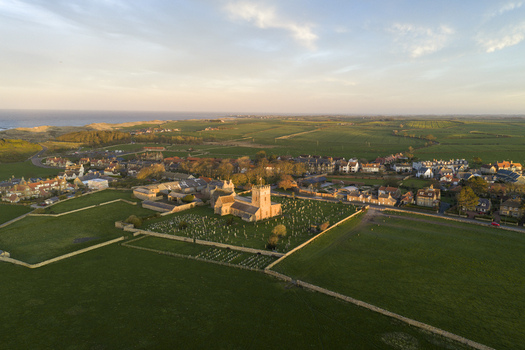 Aerial view of St Aidan s Church and its graveyard surrounded by green fields and the coastal village during golden hour Bamburgh England United Kingdom. Aerial view of St Aidan s Church and its graveyard surrounded by green fields and the coastal village during golden hour Bamburgh England United Kingdom.