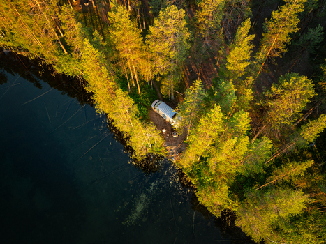 Aerial view of a campervan parked in a dense pine forest beside a dark lake during golden hour in Sweden. Aerial view of a campervan parked in a dense pine forest beside a dark lake during golden hour in Sweden.