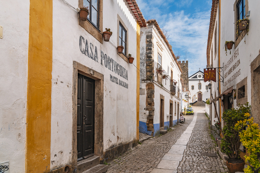 Obidos Leiria District Portugal 13 May 2026 View of a narrow cobblestone street lined with historic whitewashed buildings yellow trim potted plants and a distant church tower. Obidos Leiria District Portugal 13 May 2026 View of a narrow cobblestone street lined with historic whitewashed buildings yellow trim potted plants and a distant church tower.
