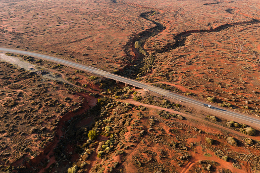 Aerial view of a long asphalt road and bridge crossing a winding dry creek bed in the vast outback landscape with a lone vehicle in Flinders Ranges South Australia Australia.