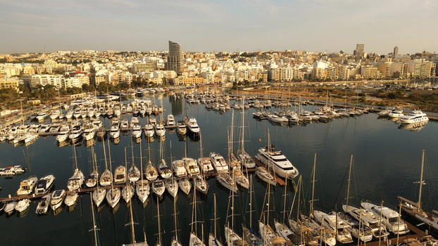 Aerial view of a marina filled with numerous yachts and sailboats docked in calm water with a dense urban cityscape and modern buildings in the background Malta. Aerial view of a marina filled with numerous yachts and sailboats docked in calm water with a dense urban cityscape and modern buildings in the background Malta.