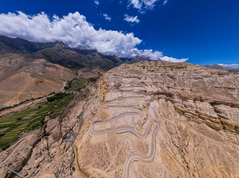 Aerial view of a winding mountain road cutting through arid rocky terrain with terraced green fields and high mountain peaks under a bright blue sky in Nepal.