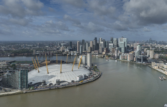 Aerial view of The O2 arena on the Greenwich Peninsula with the River Thames and the Canary Wharf skyline in the background under a cloudy sky in London England United Kingdom. Aerial view of The O2 arena on the Greenwich Peninsula with the River Thames and the Canary Wharf skyline in the background under a cloudy sky in London England United Kingdom.