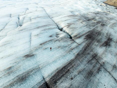 Aerial view of Breidamerkurjokull glacier with a lone hiker standing on the icy surface showcasing deep crevasses and dark volcanic ash streaks in Sveitarfelagid Hornafjordur Iceland. Aerial view of Breidamerkurjokull glacier with a lone hiker standing on the icy surface showcasing deep crevasses and dark volcanic ash streaks in Sveitarfelagid Hornafjordur Iceland.
