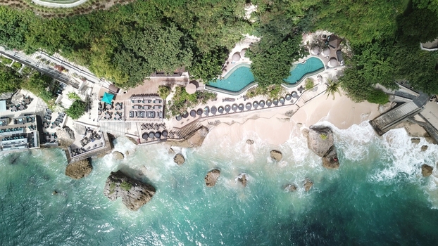 Indonesia 20 February 2026 Aerial view of a beach resort with a curved swimming pool sun loungers sandy beach and rocky coastline meeting the turquoise ocean water.