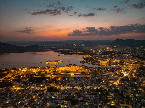 Aerial view of the illuminated City Palace and Lake Pichola at dusk with the city lights of Udaipur reflecting on the water in Udaipur Rajasthan India.