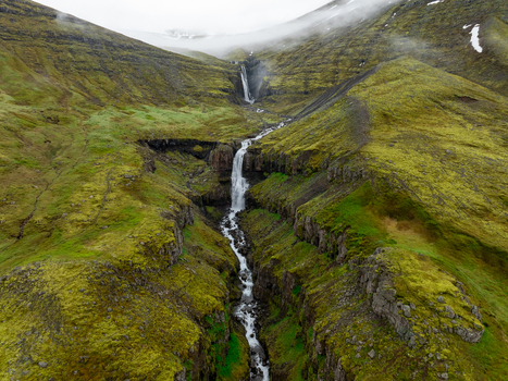 Aerial view of a cascading waterfall flowing through a lush green mossy valley in the Eastern Region of Iceland. Aerial view of a cascading waterfall flowing through a lush green mossy valley in the Eastern Region of Iceland.