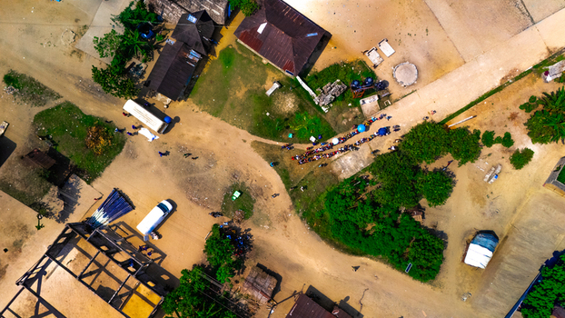 Aerial view of a rural settlement featuring small houses with dark roofs a line of people waiting along a dirt road and an unfinished building structure in Abua Rivers Nigeria. Aerial view of a rural settlement featuring small houses with dark roofs a line of people waiting along a dirt road and an unfinished building structure in Abua Rivers Nigeria.