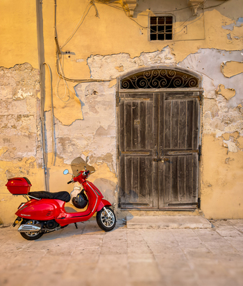 View of a vibrant red scooter parked against a weathered yellow wall featuring a rustic ornate wooden door in Valletta Malta. View of a vibrant red scooter parked against a weathered yellow wall featuring a rustic ornate wooden door in Valletta Malta.