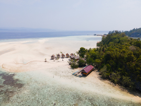 Aerial view of Ransiwor homestay with wooden huts on a white sand beach surrounded by turquoise water coral reefs and lush tropical vegetation in Raja Ampat Southwest Papua Indonesia. Aerial view of Ransiwor homestay with wooden huts on a white sand beach surrounded by turquoise water coral reefs and lush tropical vegetation in Raja Ampat Southwest Papua Indonesia.