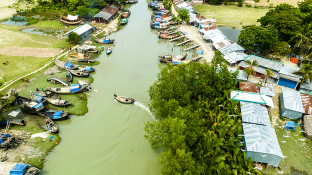 Aerial view of Patharghata featuring numerous wooden fishing boats moored along the riverbank with small houses and lush vegetation in Patharghata Barisal Division Bangladesh. Aerial view of Patharghata featuring numerous wooden fishing boats moored along the riverbank with small houses and lush vegetation in Patharghata Barisal Division Bangladesh.