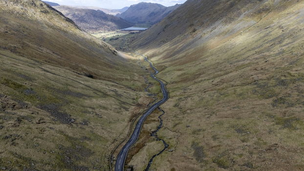 Aerial view of a winding road slicing through the rugged sun kissed valley nestled between towering peaks in a scene of natural grandeur Lake District England United Kingdom. Aerial view of a winding road slicing through the rugged sun kissed valley nestled between towering peaks in a scene of natural grandeur Lake District England United Kingdom.