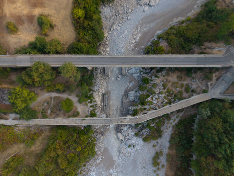 Aerial view of the historic Mesi Bridge spanning a dry rocky riverbed with a modern road bridge nearby and lush green trees in Boks Qarku i Shkodres Albania.