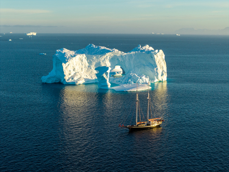 Aerial view of a massive arch iceberg floating in the deep blue waters of the Arctic with a sailboat nearby under a clear sky in Greenland. Aerial view of a massive arch iceberg floating in the deep blue waters of the Arctic with a sailboat nearby under a clear sky in Greenland.
