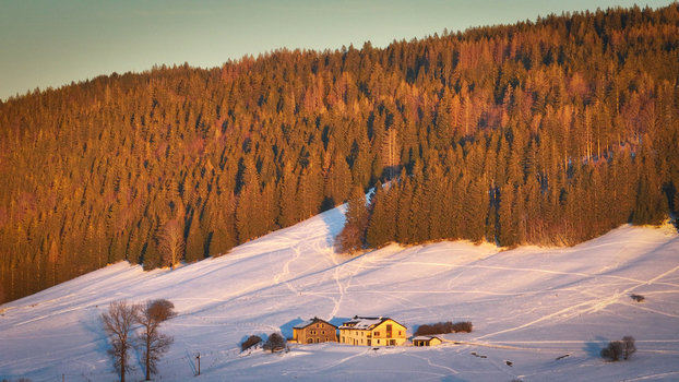 Aerial view of a snow covered landscape dotted with trails leading to a cluster of buildings nestled at the foot of a dense sun kissed forest Les Rousses Bourgogne Franche Comté France. Aerial view of a snow covered landscape dotted with trails leading to a cluster of buildings nestled at the foot of a dense sun kissed forest Les Rousses Bourgogne Franche Comté France.