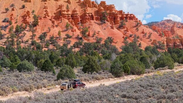 Aerial view of an SUV pulling a teardrop camper trailer along a dirt road through the red rock formations and pine trees of Costa Canyon Panguitch Utah United States.