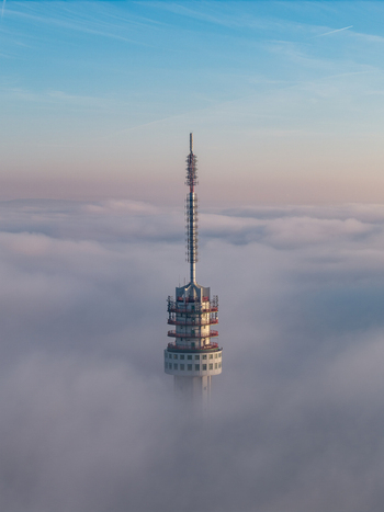 Aerial view of a tall telecommunications tower rising above a dense layer of white clouds under a clear blue sky in Roermond Limburg Netherlands.