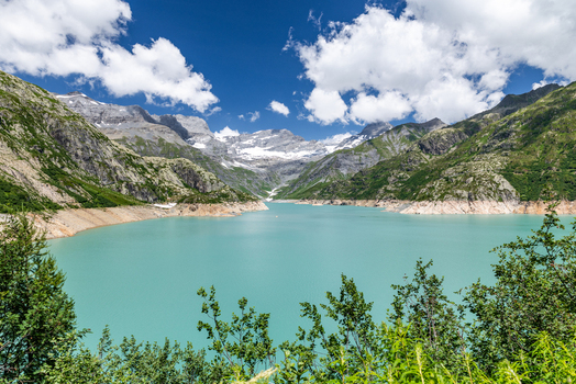 View of the turquoise waters of Lac d Emosson surrounded by snow capped peaks with green vegetation in the foreground under a bright blue sky in Le Chatelard Fribourg Switzerland. View of the turquoise waters of Lac d Emosson surrounded by snow capped peaks with green vegetation in the foreground under a bright blue sky in Le Chatelard Fribourg Switzerland.