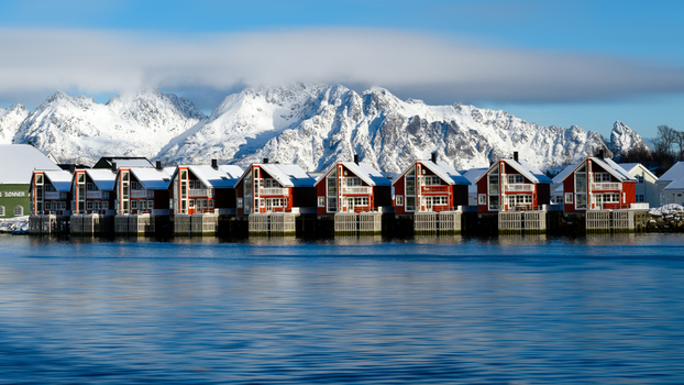 View of traditional red wooden rorbuer houses on stilts along the waterfront with snow capped mountains in the background in Leknes Lofoten Norway.