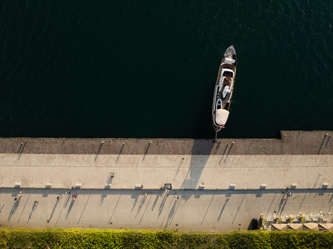 Aerial view of a luxury yacht docked at the Island of Ortigia promenade with people walking and casting long shadows in Syracuse Sicily Italy. Aerial view of a luxury yacht docked at the Island of Ortigia promenade with people walking and casting long shadows in Syracuse Sicily Italy.