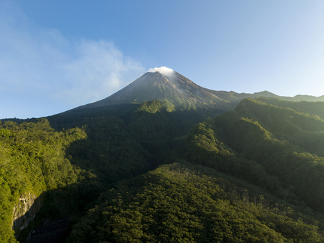 Aerial view of Mount Merapi volcano with a smoking peak and lush green forested slopes under a clear blue sky in Kecamatan Pakem Daerah Istimewa Yogya Indonesia. Aerial view of Mount Merapi volcano with a smoking peak and lush green forested slopes under a clear blue sky in Kecamatan Pakem Daerah Istimewa Yogya Indonesia.
