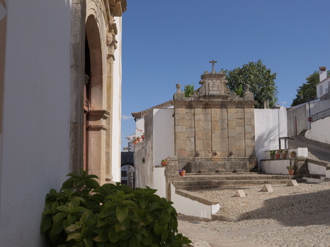 View of a historic stone fountain with a cross in a cobblestone square surrounded by traditional white buildings under a clear blue sky in Marvao Portalegre District Portugal. View of a historic stone fountain with a cross in a cobblestone square surrounded by traditional white buildings under a clear blue sky in Marvao Portalegre District Portugal.