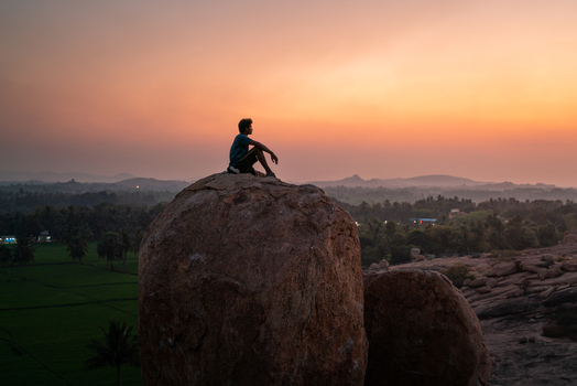 View of a man sitting on a large boulder at sunset overlooking the landscape with palm trees and fields in Hampi Karnataka India. View of a man sitting on a large boulder at sunset overlooking the landscape with palm trees and fields in Hampi Karnataka India.