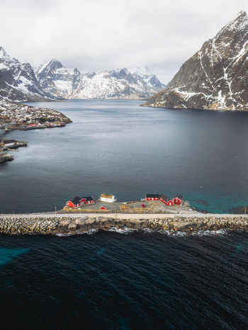 Aerial view of red cabins clustered along the rugged coastline contrasting with the deep blue sea and snow capped mountains Sakrisøya Nordland Norway. Aerial view of red cabins clustered along the rugged coastline contrasting with the deep blue sea and snow capped mountains Sakrisøya Nordland Norway.