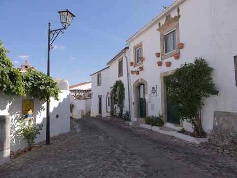 View of a historic cobblestone street in Marvao featuring traditional white washed houses with flower pots and a street lamp under a clear blue sky in Marvao Portalegre District Portugal. View of a historic cobblestone street in Marvao featuring traditional white washed houses with flower pots and a street lamp under a clear blue sky in Marvao Portalegre District Portugal.