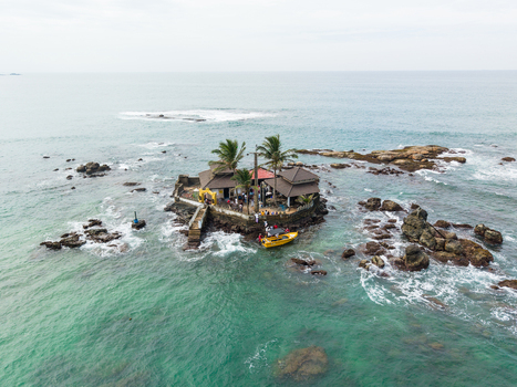 Aerial view of Seenigama Muhudu Dewalaya temple on a rocky island with palm trees and a yellow boat in turquoise waters Peraliya Sri Lanka. Aerial view of Seenigama Muhudu Dewalaya temple on a rocky island with palm trees and a yellow boat in turquoise waters Peraliya Sri Lanka.
