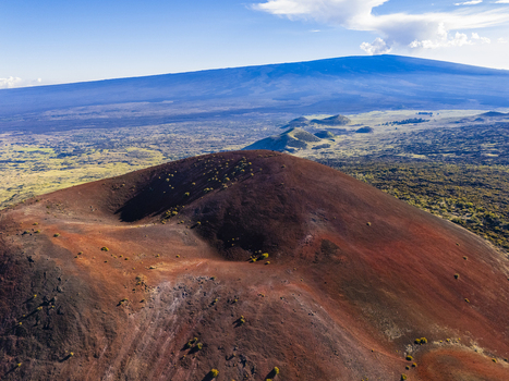 Aerial view of the reddish cinder cone of Mauna Kea volcano surrounded by vast lava fields and sparse vegetation under a clear blue sky in Hawaii County Hawaii United States.