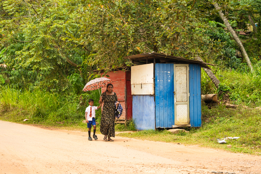 Sri Lanka 06 February 2026 View of a woman holding an umbrella and a young boy in school uniform walking along a dirt road past a small colorful shack in a rural area.