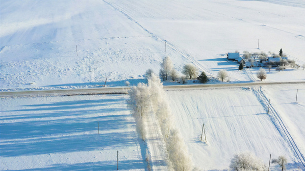Aerial view of a snow covered rural landscape with frost covered trees lining a road and a small house in the distance in Latvia. Aerial view of a snow covered rural landscape with frost covered trees lining a road and a small house in the distance in Latvia.